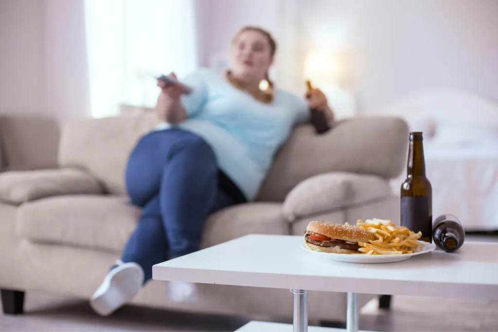 Tavolino in primo piano con junk food e ragazza sovrappeso sullo sfondo che guarda la tv