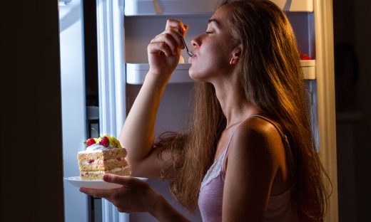 Hungry woman in pajamas enjoys sweet cake at night near refrigerator. Stop diet and gain extra pounds due to carbs food and unhealthy eating