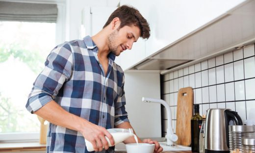 Attractive young man in plaid shirt standing and pouring milk into bowl for breakfast on the kitchen