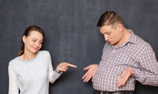 Studio waist-up shot of cheerful woman pointing with finger at big stomach of man being indignant and displeased, shrugging shoulder with bewilderment, over gray background. Man's obesity