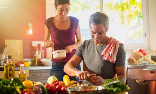 Trendy couple in casual clothes cooking vegetables from the market in a red kitchen. The woman is stirring the sauce in a white pan while the grey hair man is taking care of the fresh vegetables.Backlit shot with flare